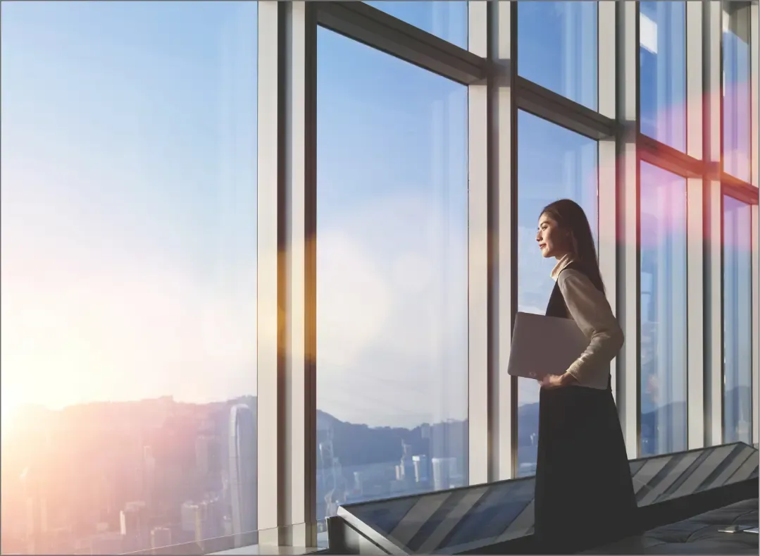A professional woman standing by large office windows holding a laptop, looking out at the sunrise with a sense of ambition and determination.